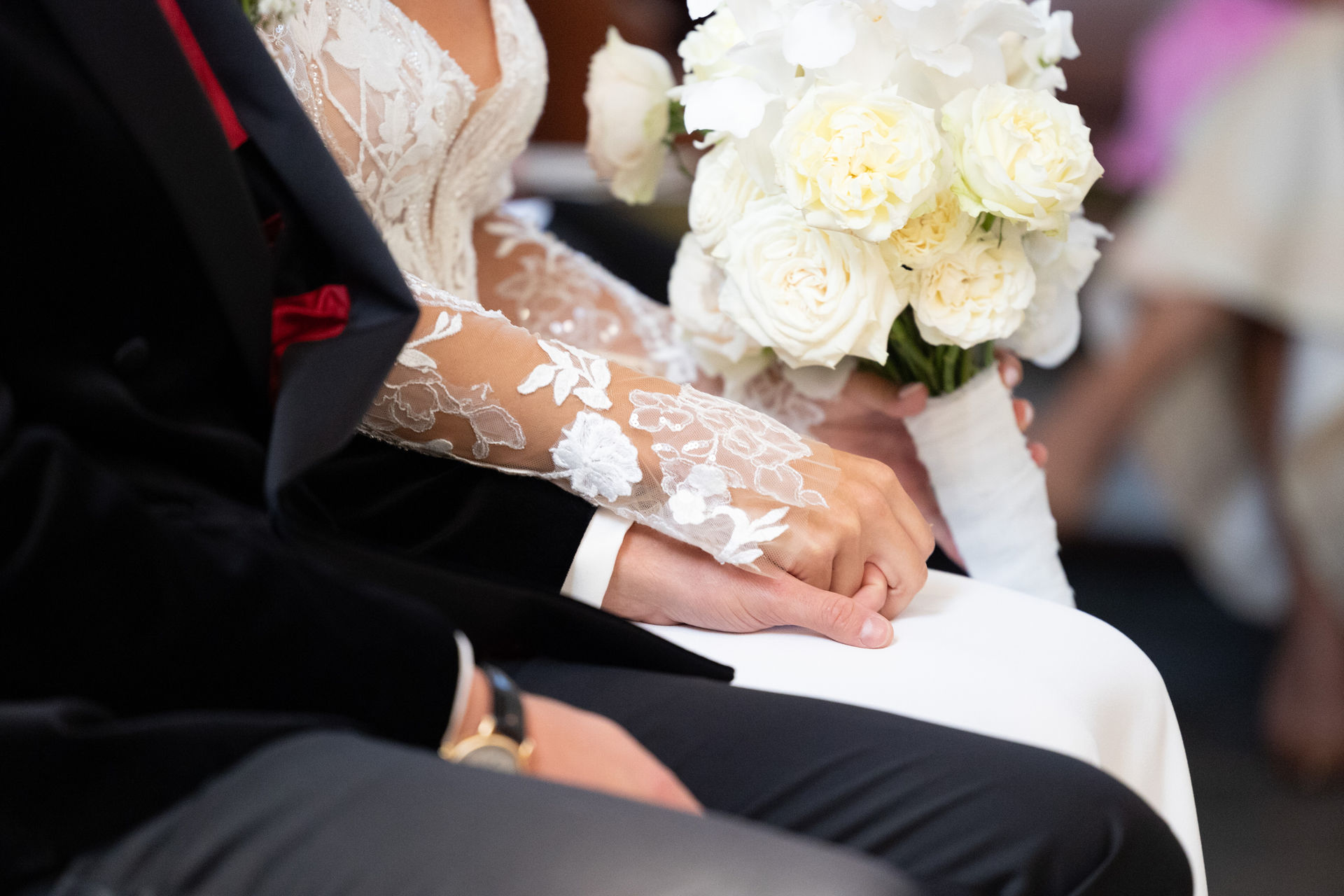 Nahaufnahme: Die Hände eines Brautpaares halten sich, die Braut trägt ein langes Ärmelkleid und hält einen Blumenstrauß.