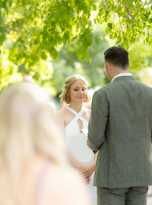 Eine Frau in weißem Kleid und ein Mann im Anzug stehen sich unter Bäumen gegenüber.