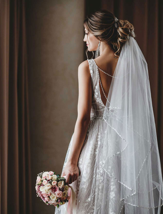 Braut in Hochzeitskleid mit Schleier und Blumenstrauß, von hinten fotografiert, schaut zur Seite.