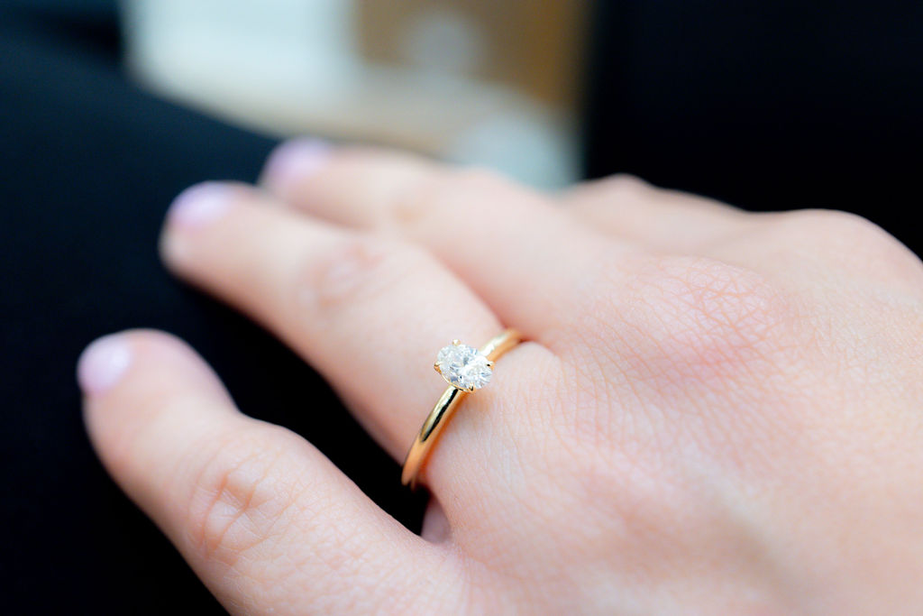 Eine Hand mit einem goldenen Ring an einem Finger. Der Ring hat einen einzelnen, ovalen, weißen Stein.