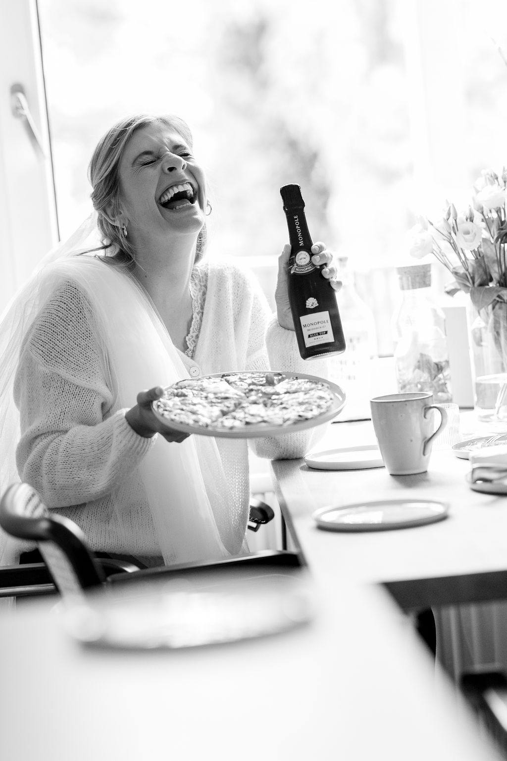 Eine lachende Frau mit hautfarbenem Pullover hält eine Pizza und eine Flasche in ihren Händen.