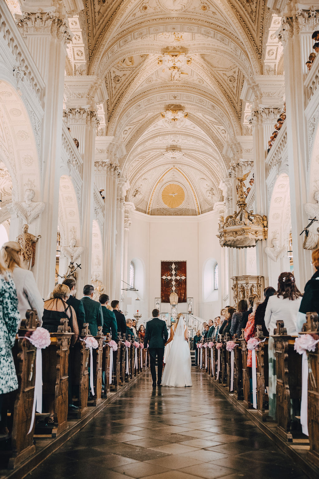 Ein Brautpaar geht einen Mittelgang in einer großen, reich verzierten Kirche entlang, gesäumt von Gästen.