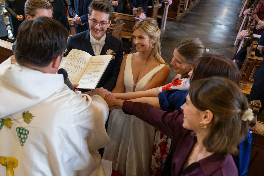 Ein Priester liest aus einem Buch, während um ihn herum eine Hochzeitsgesellschaft ihre Hände zusammenlegt.