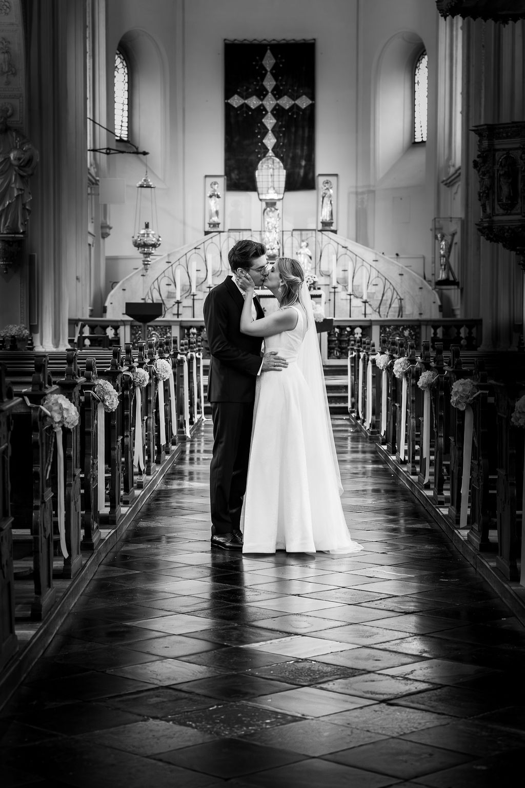 Ein Brautpaar küsst sich in einer Kirche vor dem Altarraum, zwischen dekorierten Kirchenbänken.