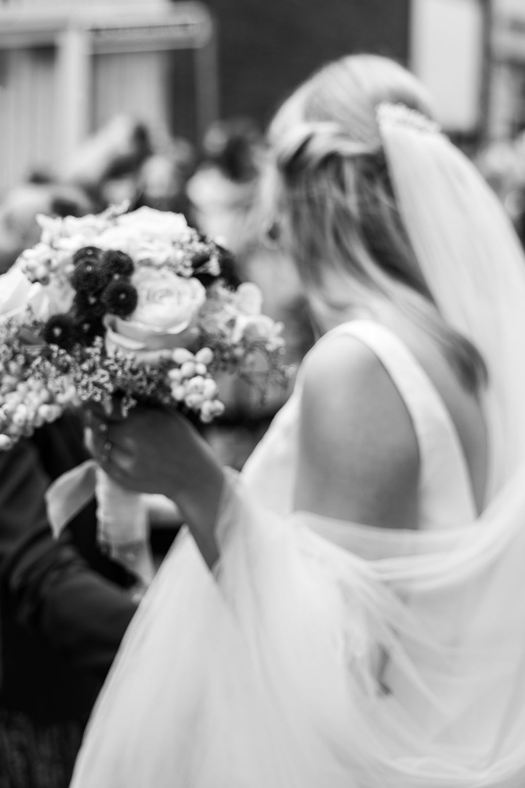 Rückenansicht einer Braut im Hochzeitskleid mit Schleier und Brautstrauß in schwarz-weiß.