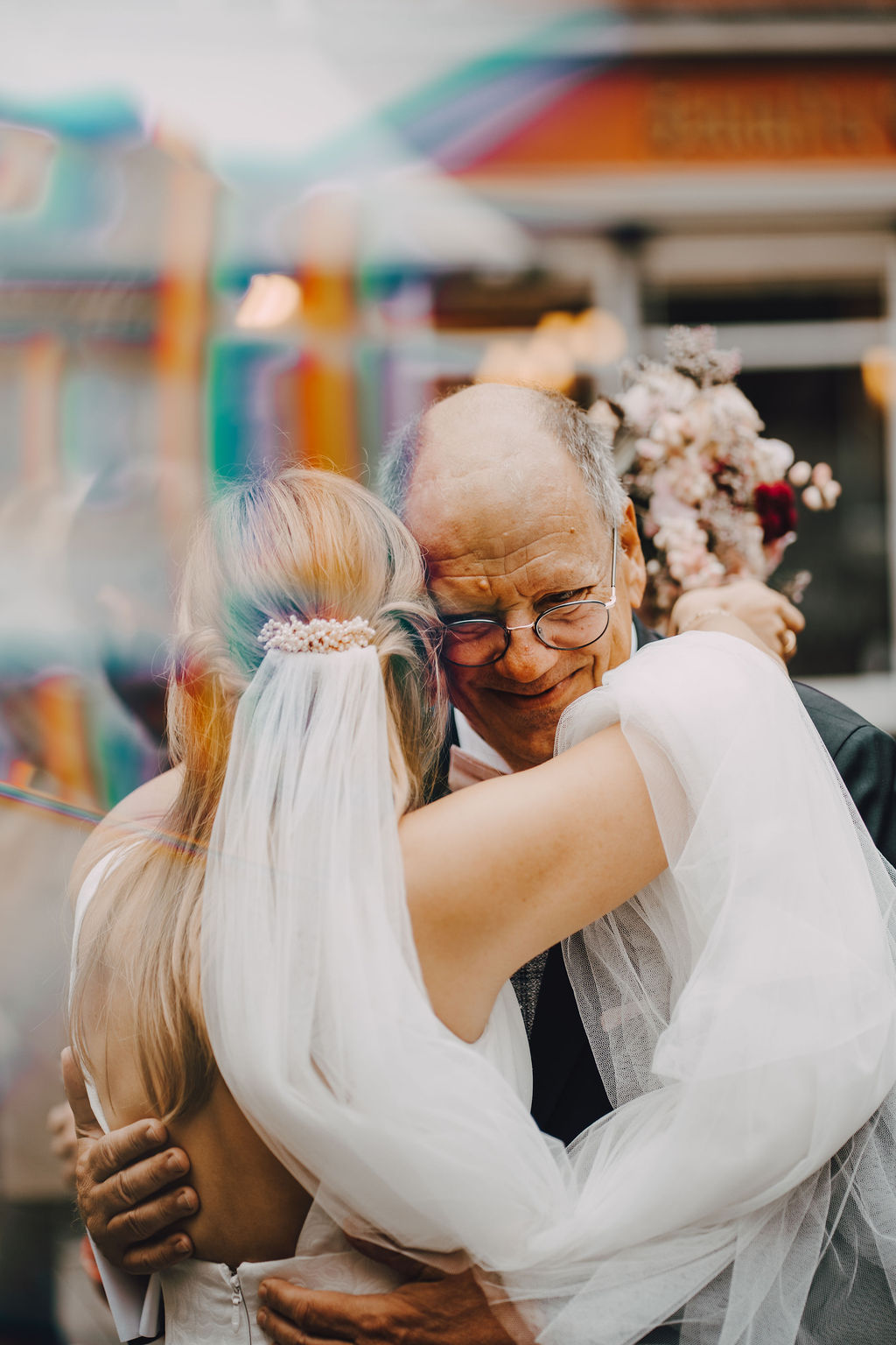 Eine Person in einem weißen Hochzeitskleid umarmt eine ältere Person mit Brille und Anzug.