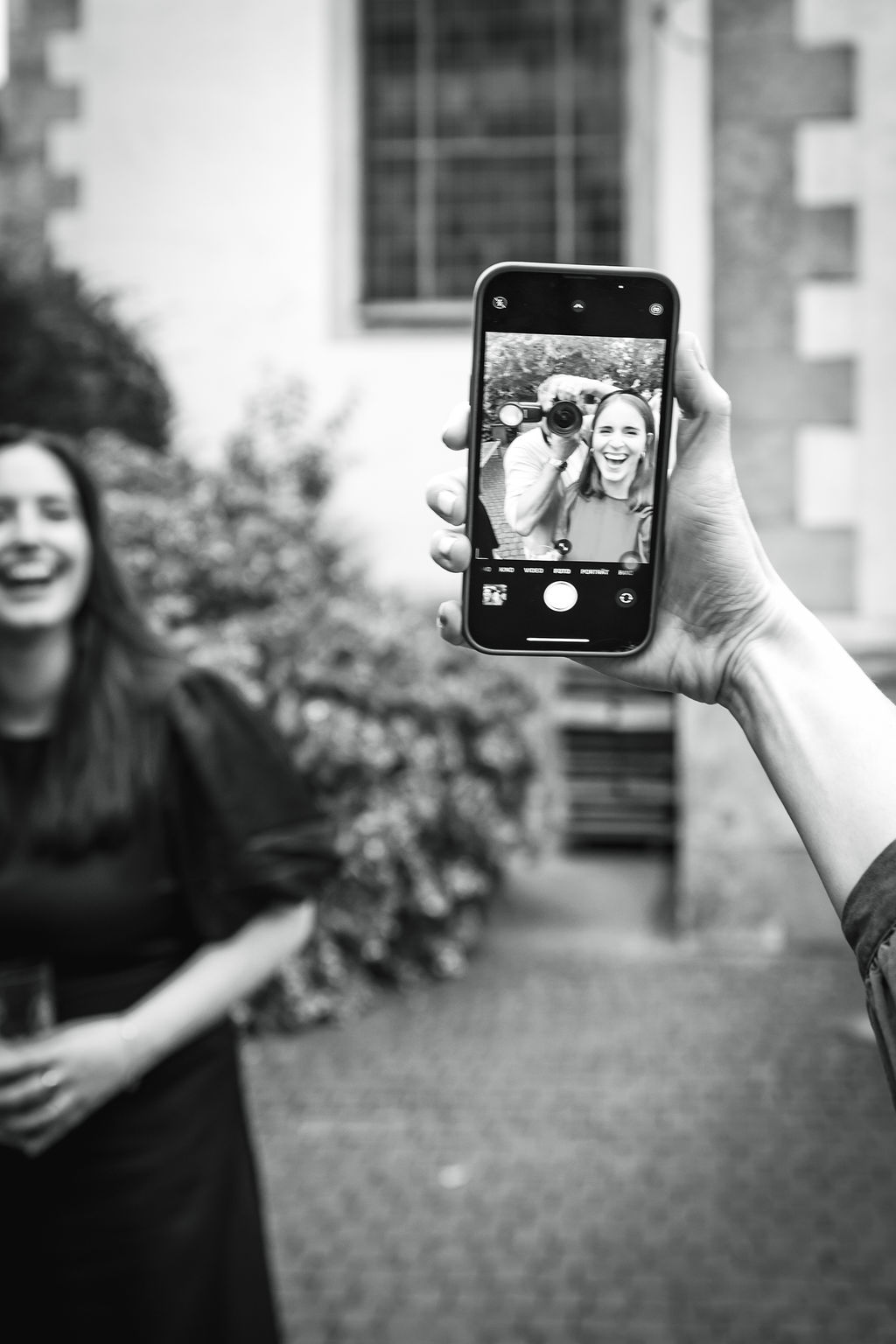 Eine Hand hält ein Smartphone mit einem lachenden Paar im Display, eine Person im Vordergrund ist unscharf.