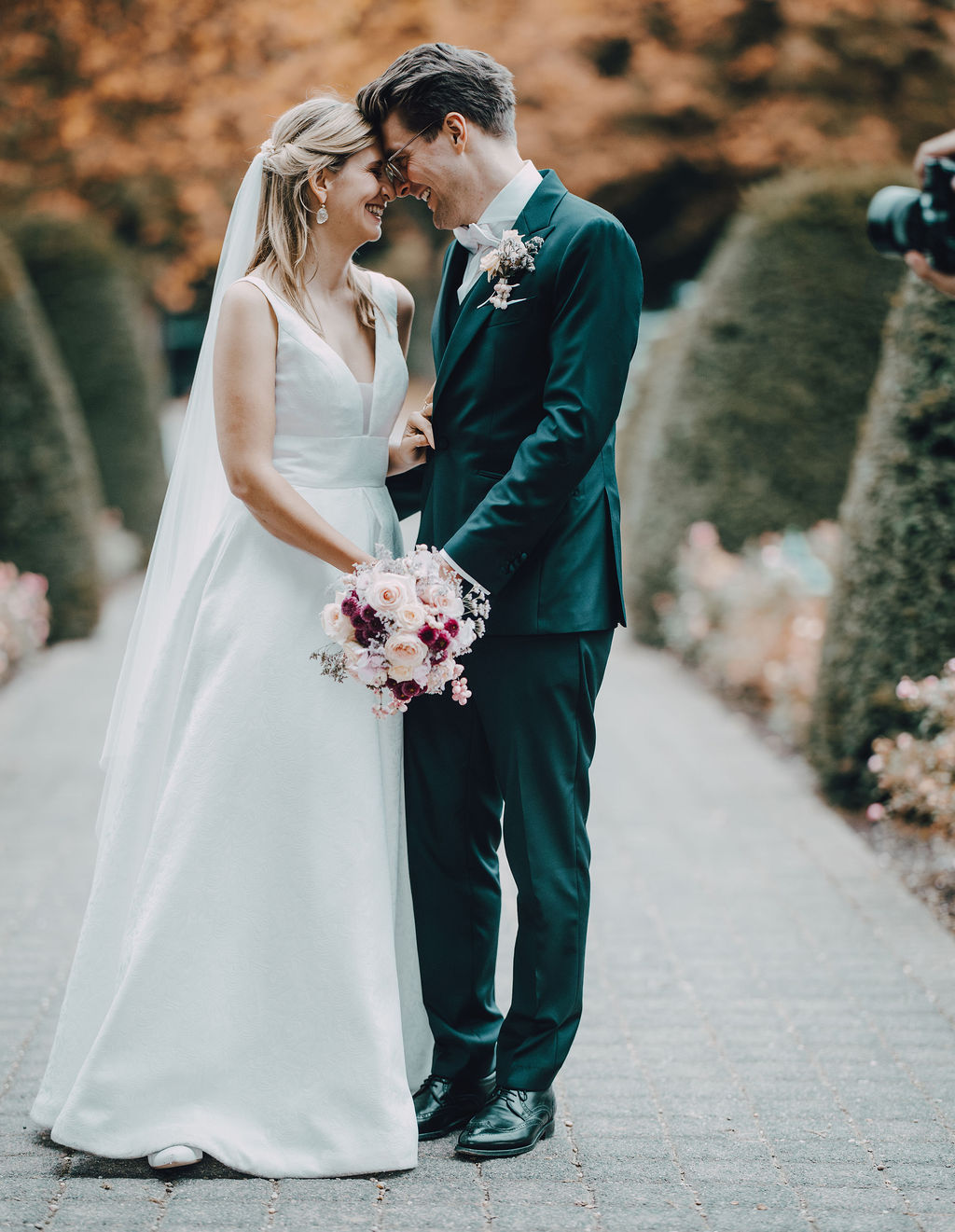 Brautpaar in Hochzeitskleidung hält Händchen und steht auf einem gepflasterten Weg in einem Garten mit Formschnitthecken.