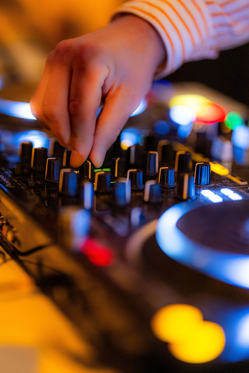 Eine Hand bedient Regler und Knöpfe an einem DJ-Mischpult mit bunten Lichtern im Hintergrund.