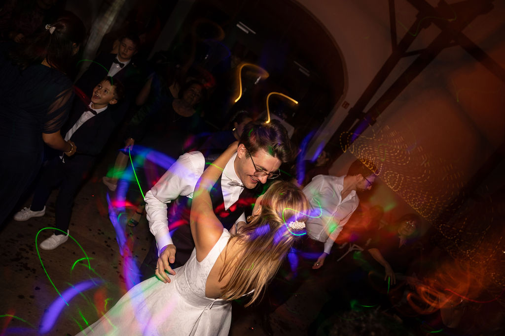 Ein Paar tanzt auf einer Hochzeit, umgeben von bunten Lichtern und weiteren Gästen. Beide lächeln.