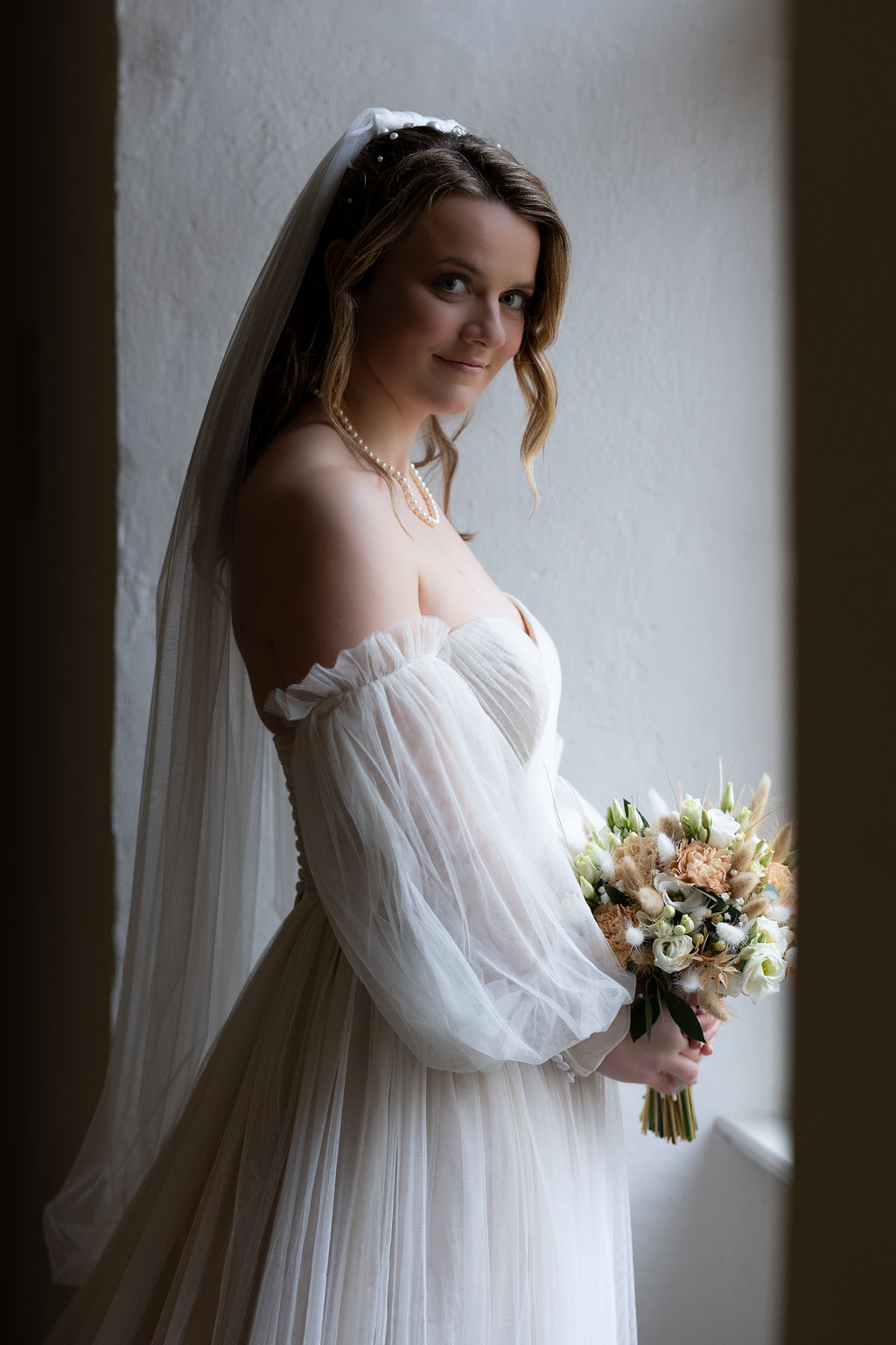 Eine Frau im Hochzeitskleid mit Schleier und Blumenstrauß steht in einem Raum, seitliches Licht.