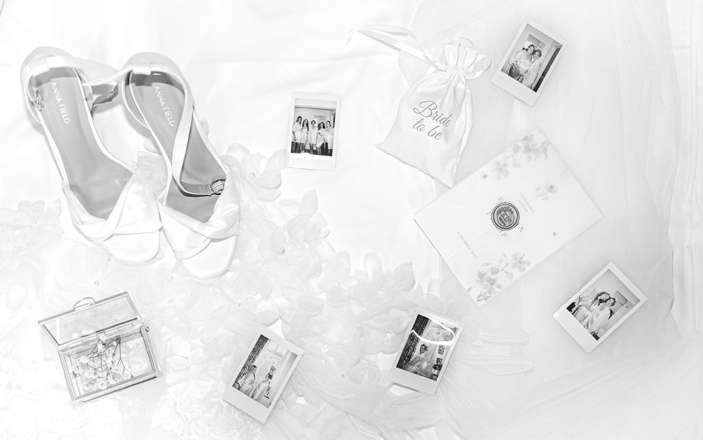 Hochzeitsaccessoires: weiße Schuhe, Beutel "Bride to be", Einladung, Glasschachtel und Fotos auf hellem Stoff.