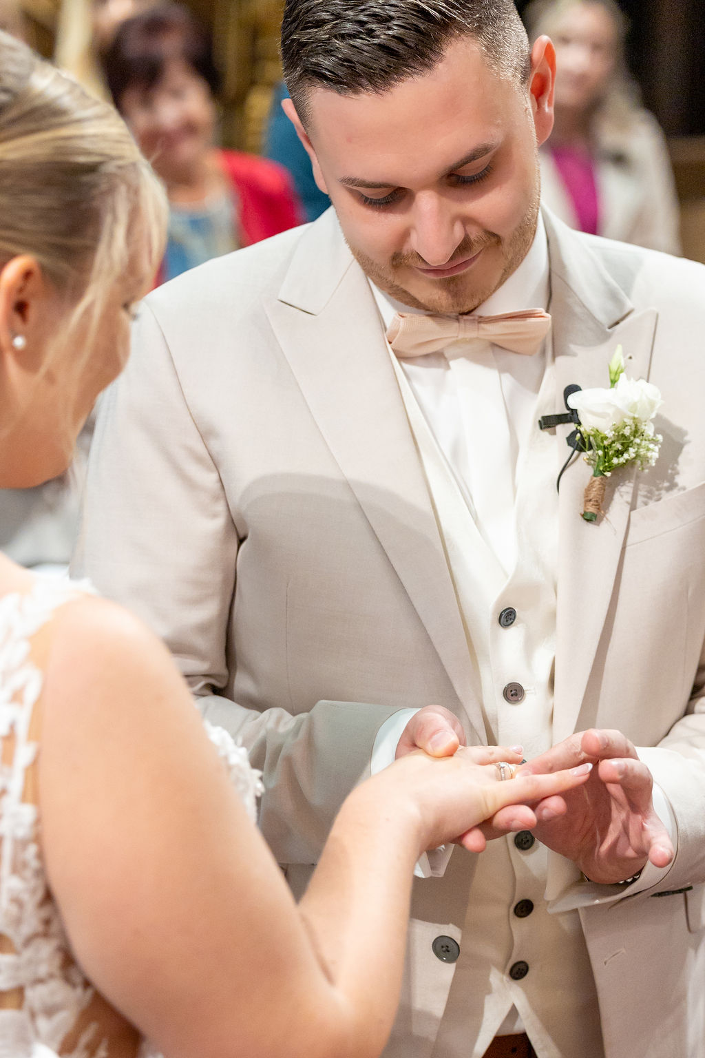 Ein Bräutigam im hellen Anzug steckt einer Braut einen Ring an. Die Braut trägt ein weißes Spitzenkleid.
