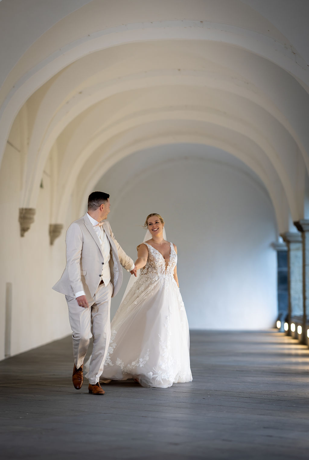 Ein Brautpaar geht Hand in Hand durch einen hellen Arkadengang. Sie trägt ein weißes Hochzeitskleid, er einen beigen Anzug.
