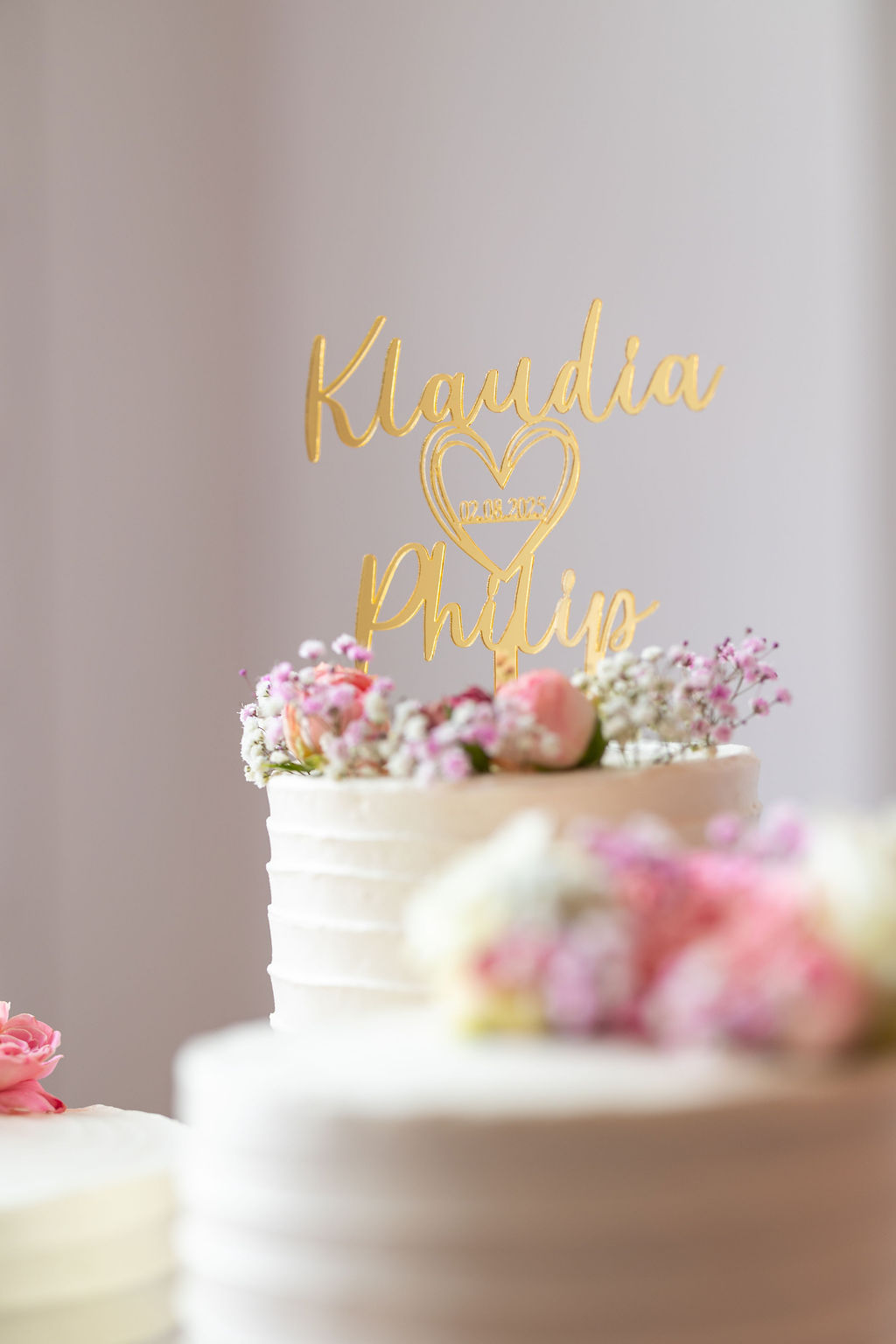 Eine goldene Tortenfigur mit den Namen "Klaudia" und "Philip" und dem Datum "02.08.2023" auf einer Hochzeitstorte.