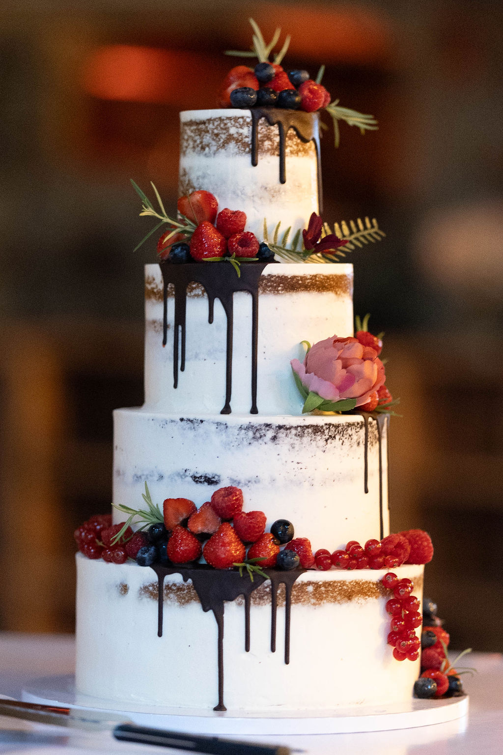 Mehrstöckige Hochzeitstorte mit Beeren, Schokodrip und einzelnen Blüten auf weißem Tortenständer.