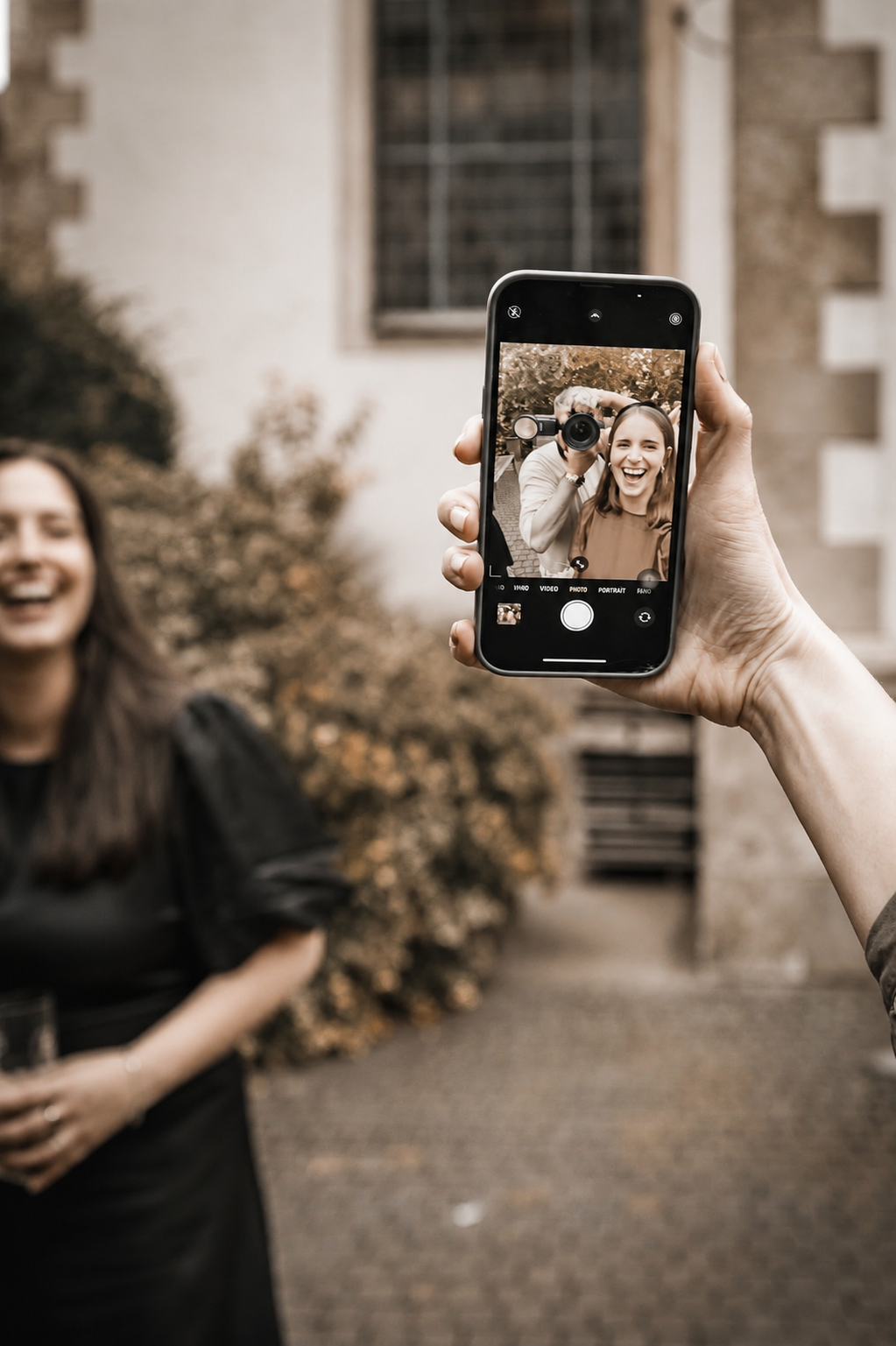 Eine Hand hält ein Smartphone mit einem Foto zweier lachender Personen, im Hintergrund unscharf eine lachende Person stehend.