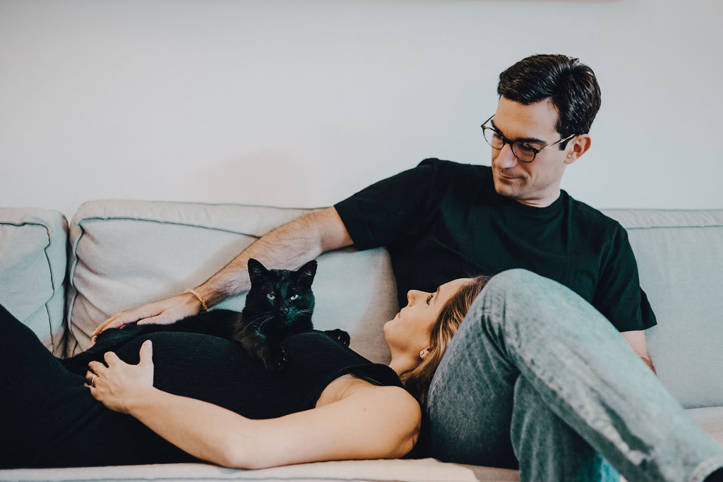 Ein Paar liegt auf einem Sofa, eine schwarze Katze liegt auf dem Bauch der Frau. Der Mann schaut die Frau an.