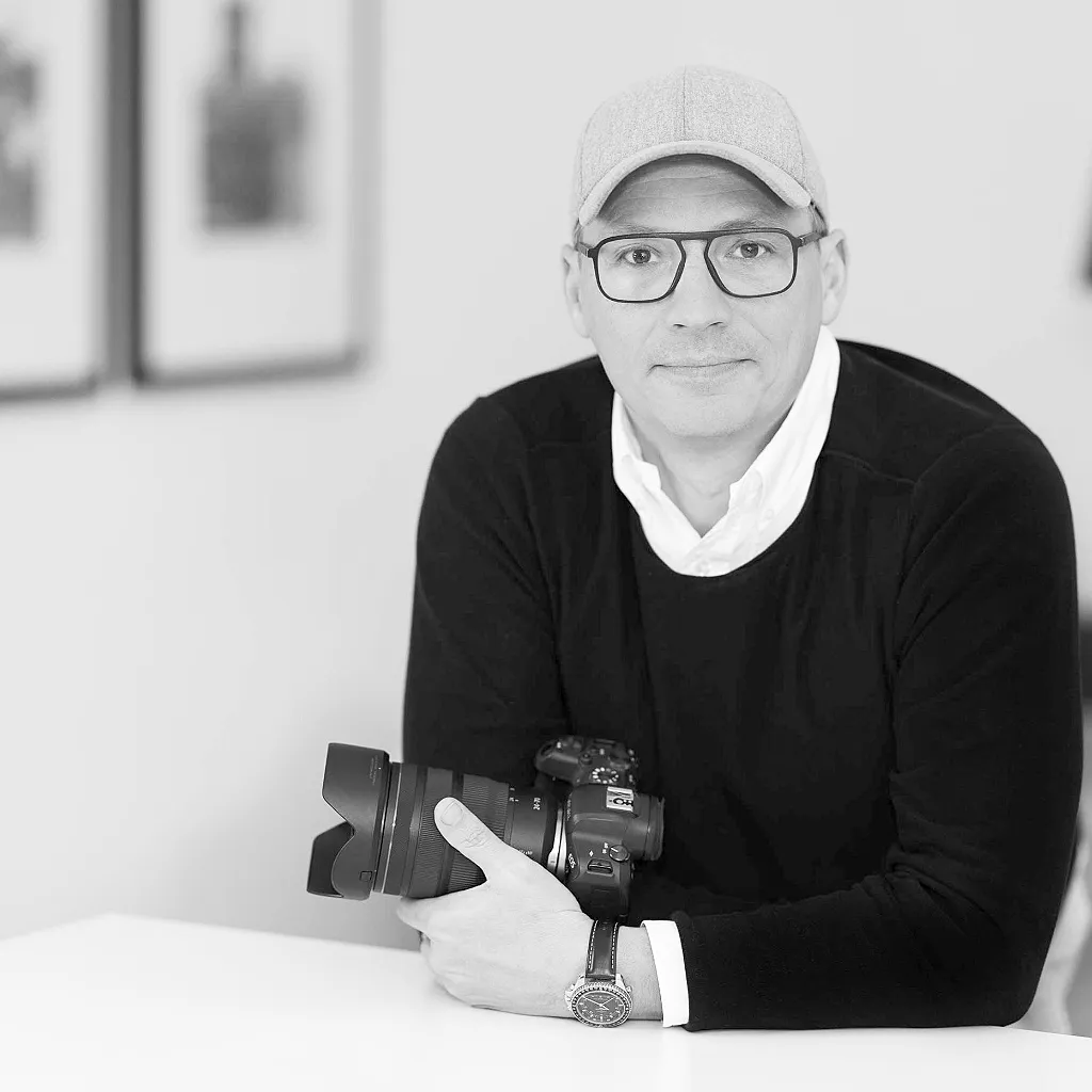 Ein Mann mit Brille, Kappe und Kamera blickt sitzend in die Kamera. Schwarz-Weiß-Aufnahme.