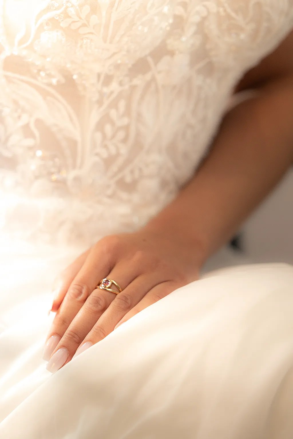 Eine goldene Hand mit Ring auf weißem Stoff vor einem mit Spitze bestickten hochzeitskleid.