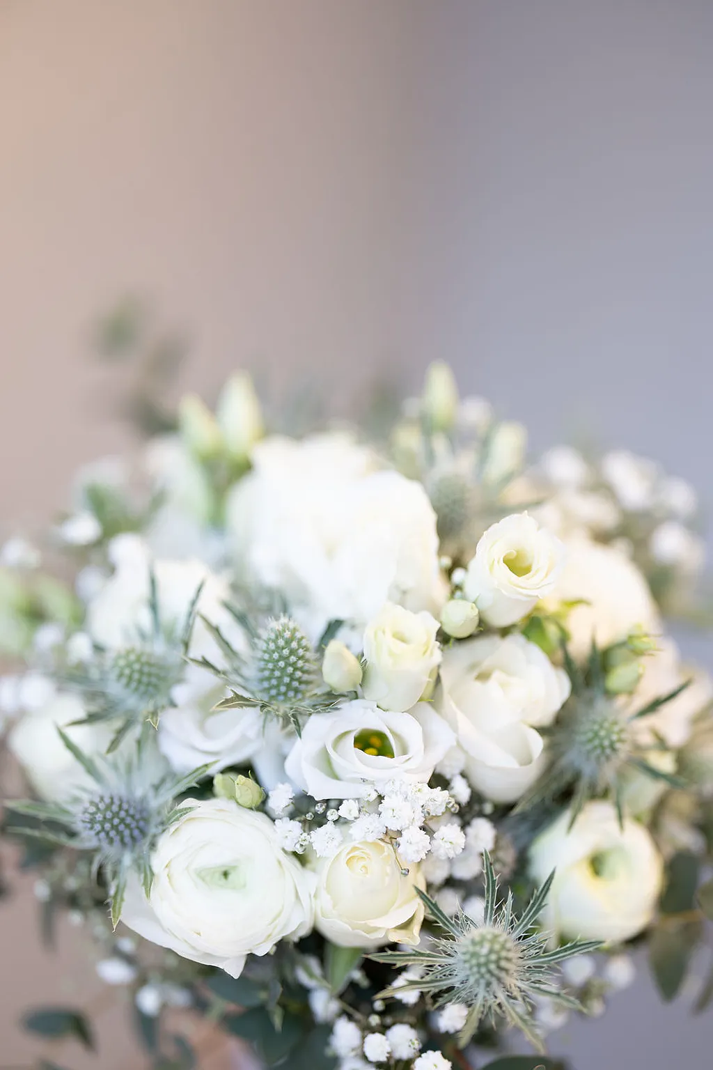 Nahaufnahme eines Brautstraußes mit weißen Rosen, Ranunkeln, Schleierkraut und Disteln vor unscharfem Hintergrund.