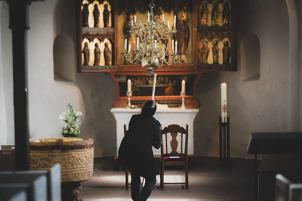 Eine Person in einer Kirche zündet eine Kerze mit einem Stab an einem Kronleuchter an.
