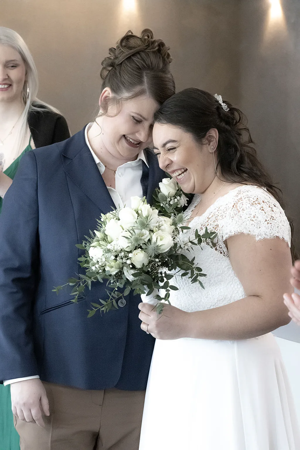 Zwei Frauen stehen eng beieinander, eine trägt einen Anzug, die andere ein weißes Kleid und hält einen Blumenstrauß.