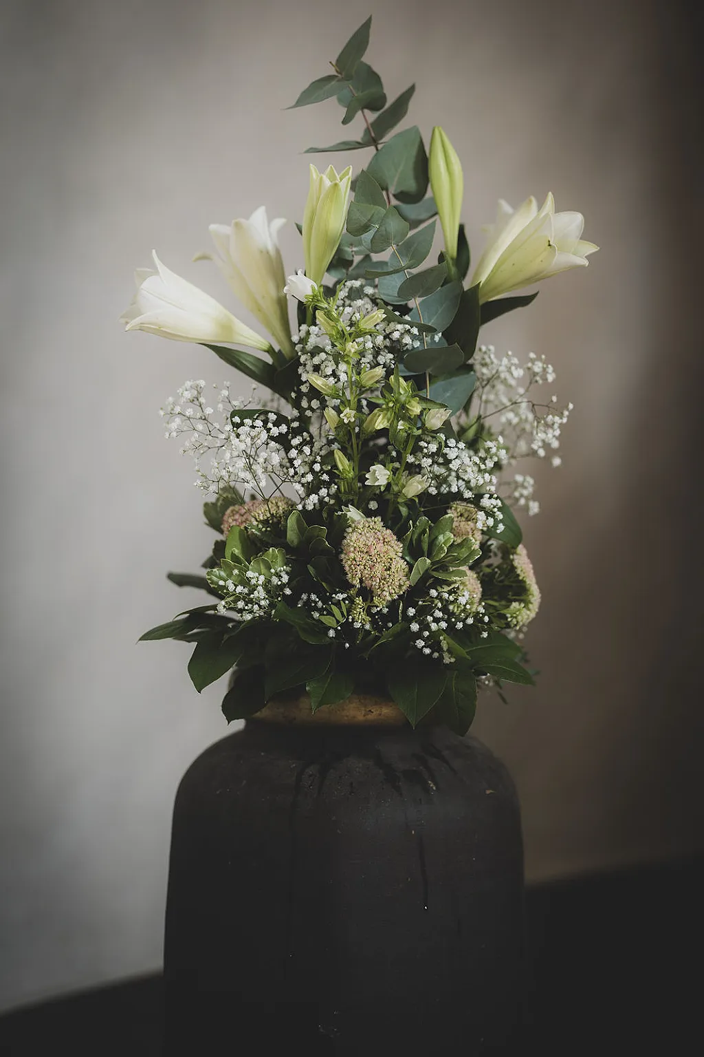 Ein Blumenarrangement mit weißen Lilien und Schleierkraut in einer dunklen Bodenvase.