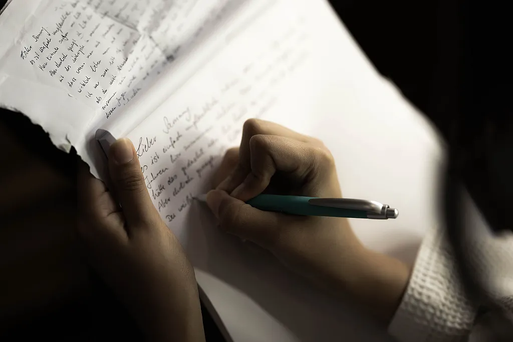 Eine Person schreibt mit einem türkisen Stift auf ein weißes Blatt Papier mit handschriftlicher Notiz.