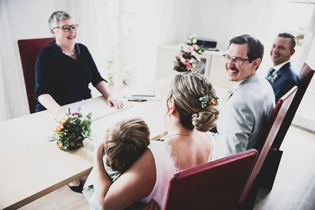 Vier Personen sitzen an einem Tisch, eine Person links lächelt, im Vordergrund ein Blumenstrauß.