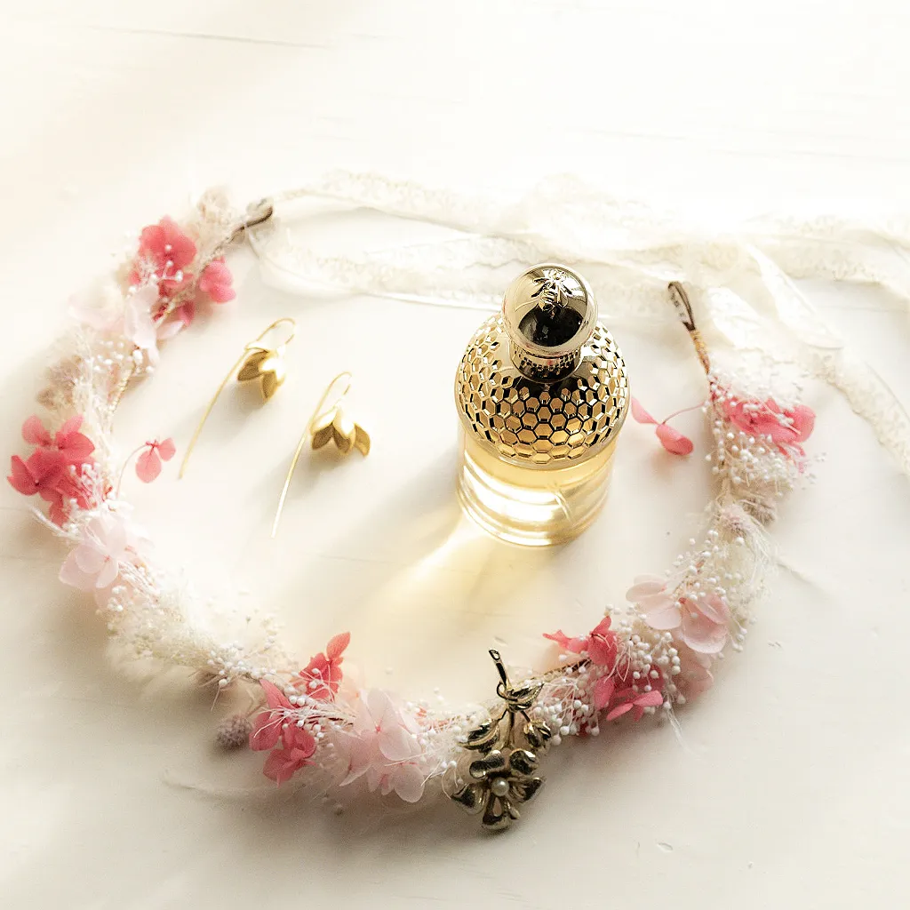 Ein Haarkranz aus rosa Blumen und kleinen weißen Blüten, goldener Schmuck und ein Parfümflakon.