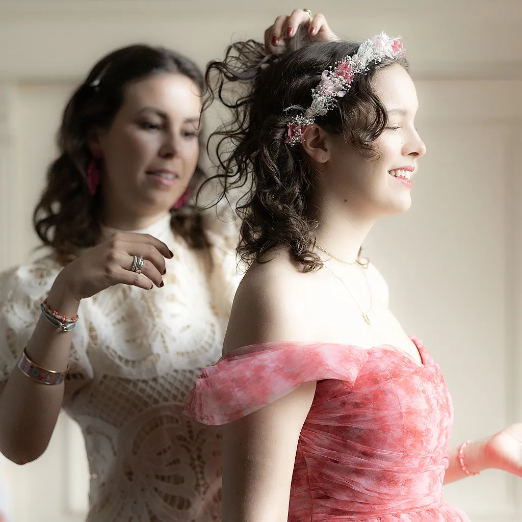 Eine Person in einem weißen Spitzenoberteil stylt die Haare einer anderen Person, die einen Blumenhaarreif und ein rosa…