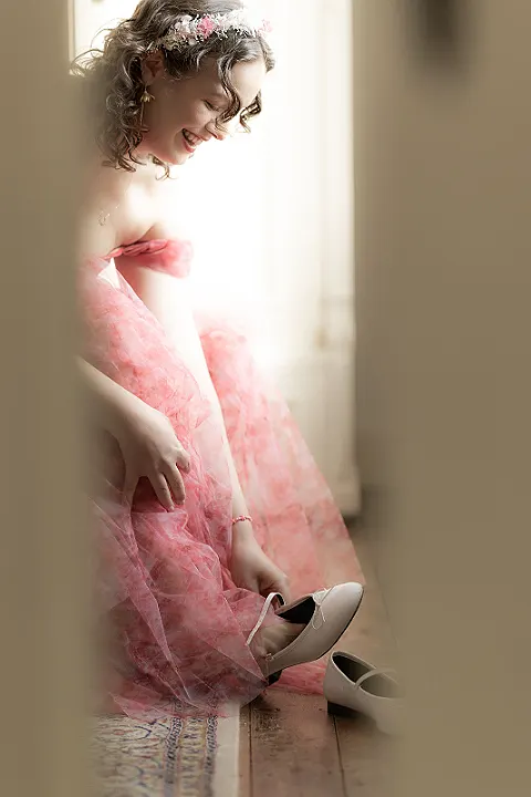 Eine Frau mit rosa Kleid und Blumen im Haar zieht sich weiße Schuhe an.