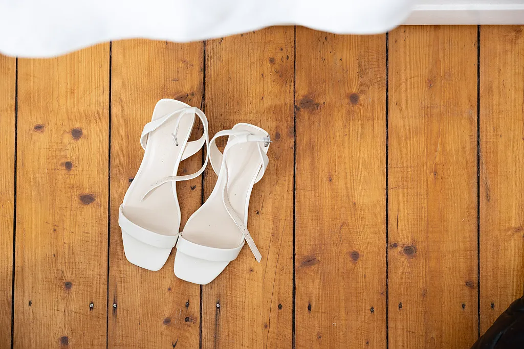 Ein Paar weiße Brautschuhe mit Riemchen stehen auf einem Holzboden. Darüber fällt ein weißer Stoff.