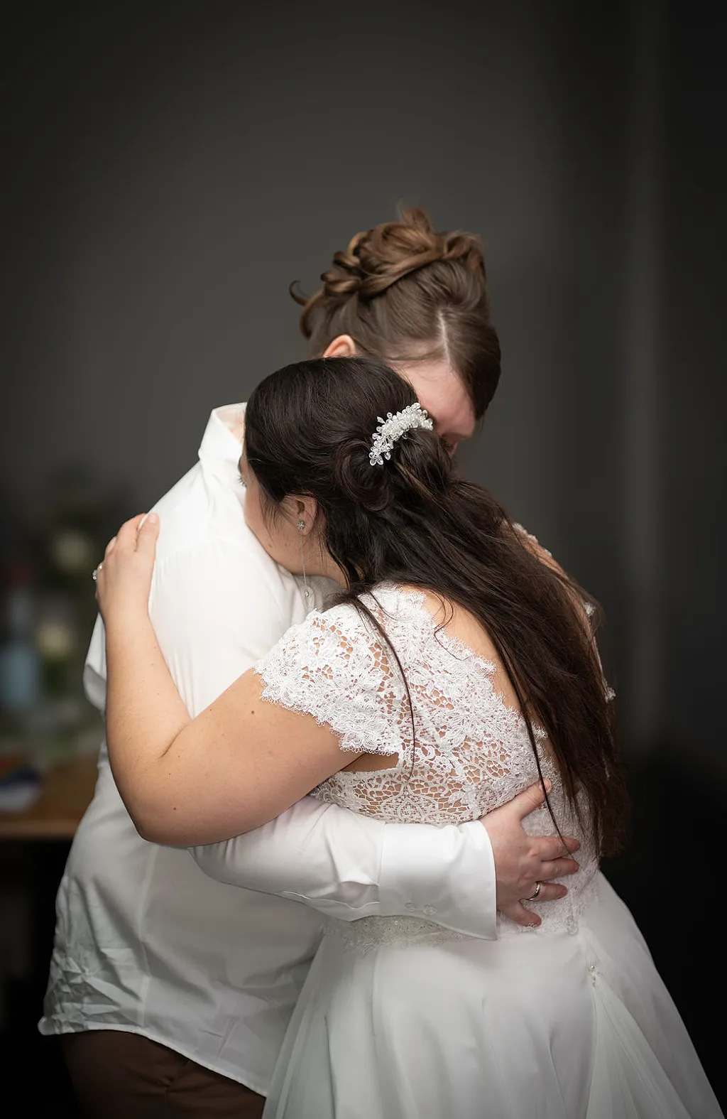 Zwei Frauen umarmen sich, eine trägt ein weißes Spitzenkleid, die andere ein weißes Hemd.