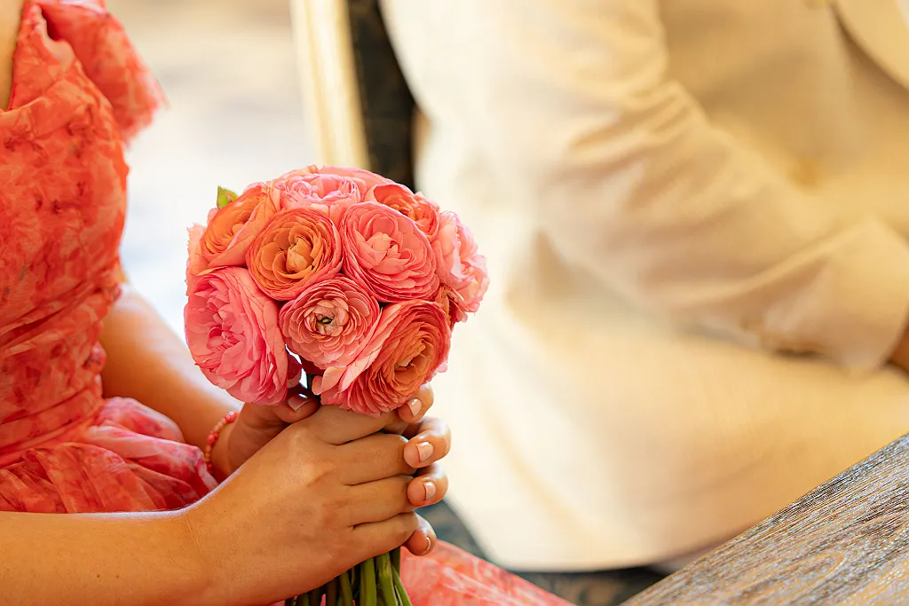 Eine Person in rotem Kleid hält einen pinkfarbenen Hochzeitsstrauß aus Ranunkeln, im Hintergrund unscharf eine zweite Person.