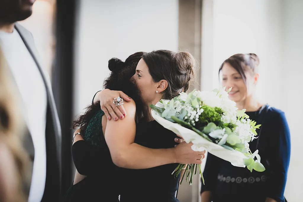 Zwei Frauen umarmen sich, eine davon hält einen weißen Blumenstrauß. Im Hintergrund weitere Personen.