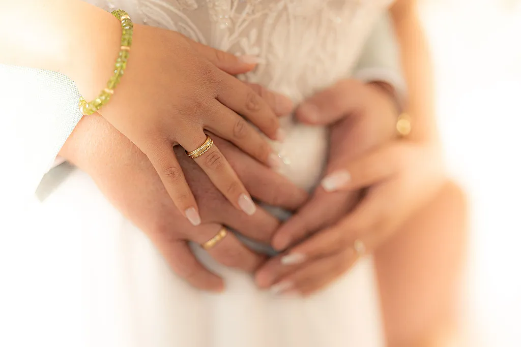 Hände eines Brautpaares, ineinander verschlungen, goldene Ringe sichtbar. Eine Hand trägt ein grünes Armband.