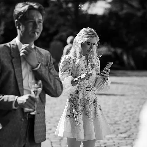 Eine Frau steht mit einem Glas Sekt und einem Smartphone in den Händen, ein Mann mit Zigarette im Mund steht daneben.