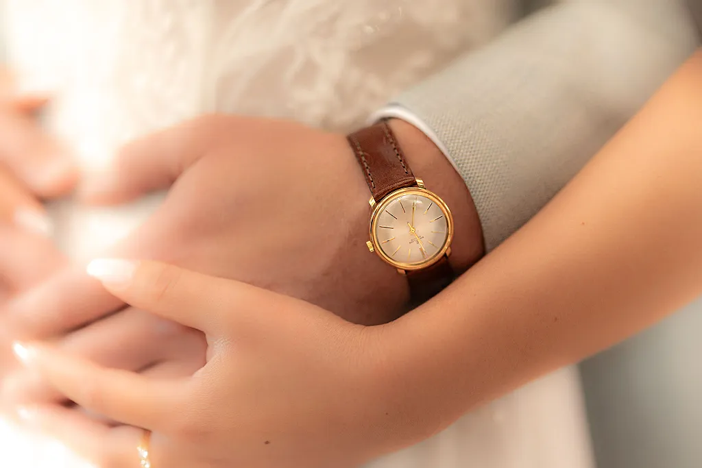 Nahaufnahme: Eine goldene Armbanduhr an einem Handgelenk mit braunem Lederarmband, umgeben von zwei Händen.