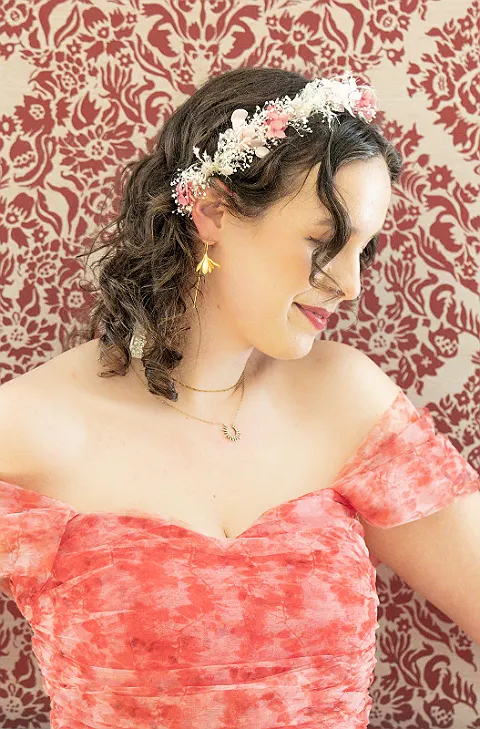 Eine Frau mit Locken, Blumen im Haar, Ohrringen und einer Halskette. Hintergrund ist ein rotes Blumenmuster.