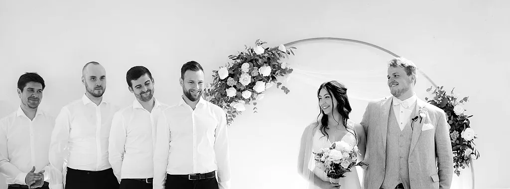 Sechs Personen vor einem weißen Hintergrund mit Blumendekoration an einem Bogen. Vier Männer links, ein Paar rechts.