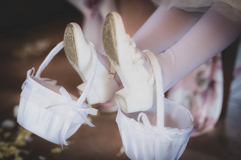 Zwei Kinderbeine in weißen Strumpfhosen und Schuhen, die jeweils einen Blumenkorb halten.