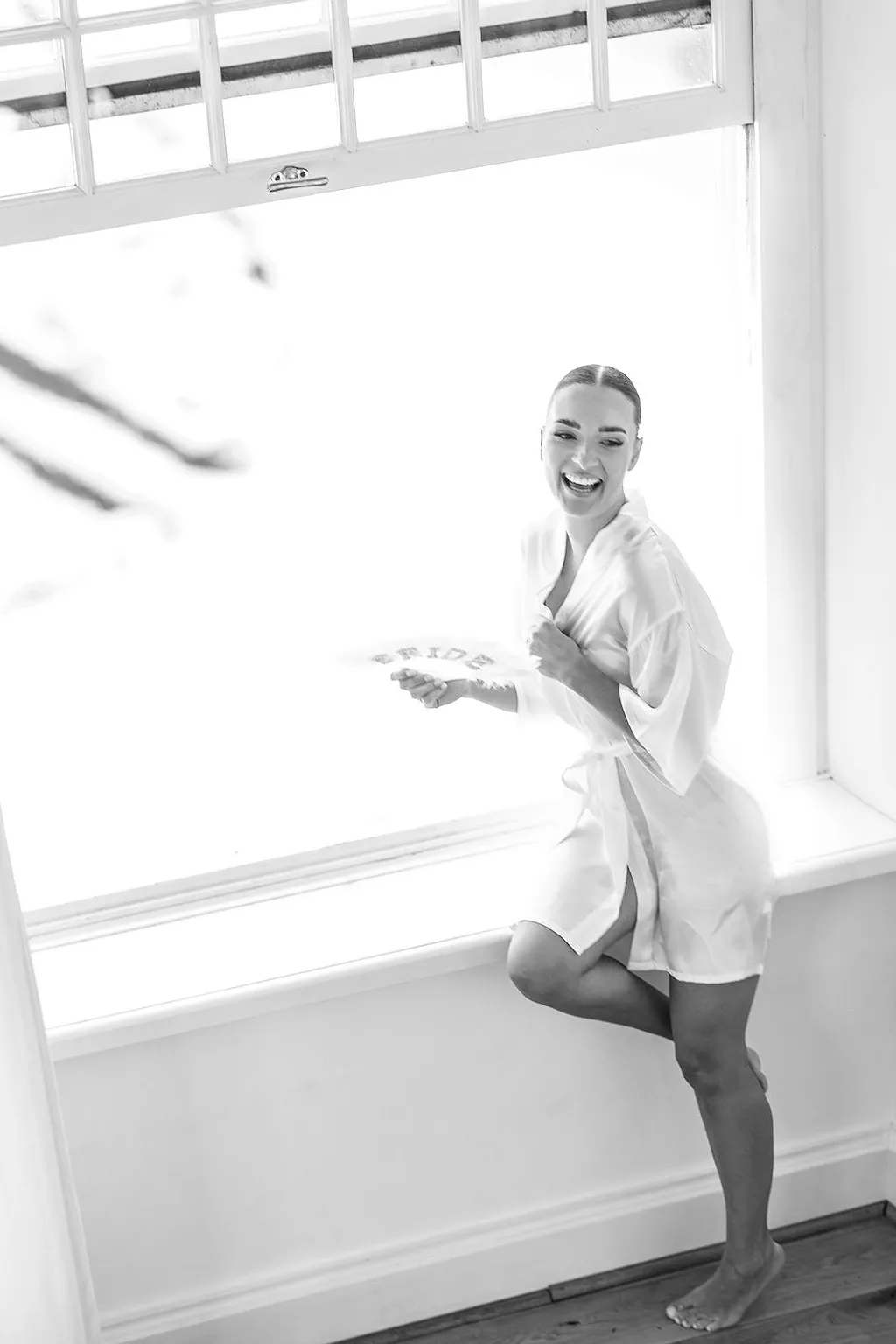 Eine lachende Frau in einem hellen Morgenmantel sitzt auf einer Fensterbank und hält einen Fächer mit der Aufschrift „Bride“.