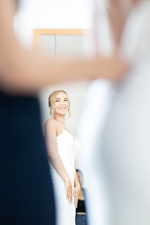 Eine lächelnde Braut in einem weißen Spaghetti-Träger-Hochzeitskleid mit einer Person im Hintergrund.