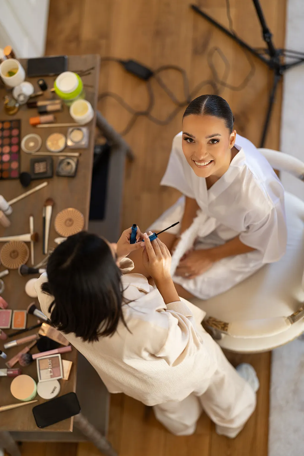 Eine Frau wird geschminkt, während Make-up-Produkte auf einem Tisch liegen und eine Lampe auf dem Boden steht.