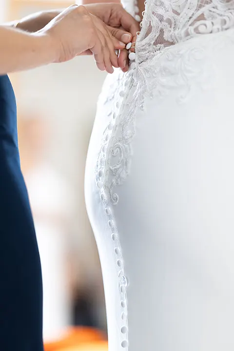 Hände schließen Knöpfe an der Rückenpartie eines weißen Brautkleides mit Spitze.