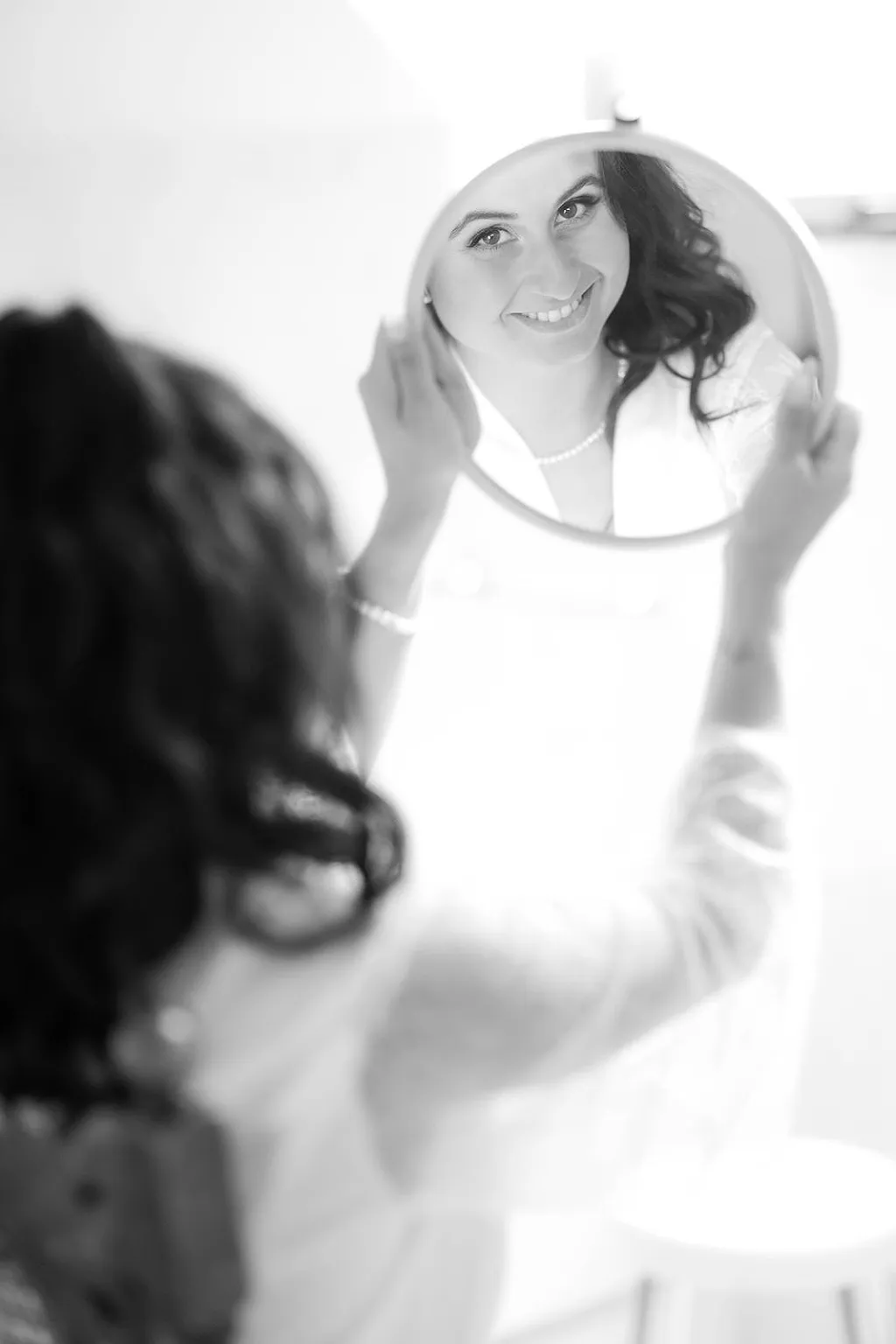 Eine Frau mit dunklen welligen Haaren betrachtet ihr Spiegelbild in einem runden Handspiegel.