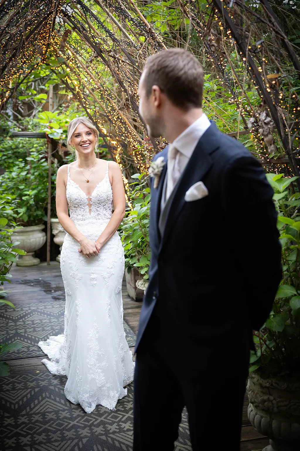 Eine Frau im Hochzeitskleid lächelt einen Mann im Anzug an; beide stehen in einem begrünten Gang mit Lichterketten.