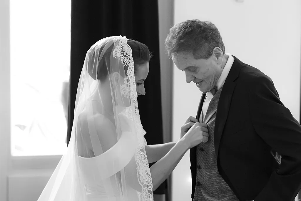 Eine Frau im Hochzeitskleid mit Schleier hilft einem Mann im Anzug beim Anziehen der Weste, Schwarz-Weiß-Aufnahme.